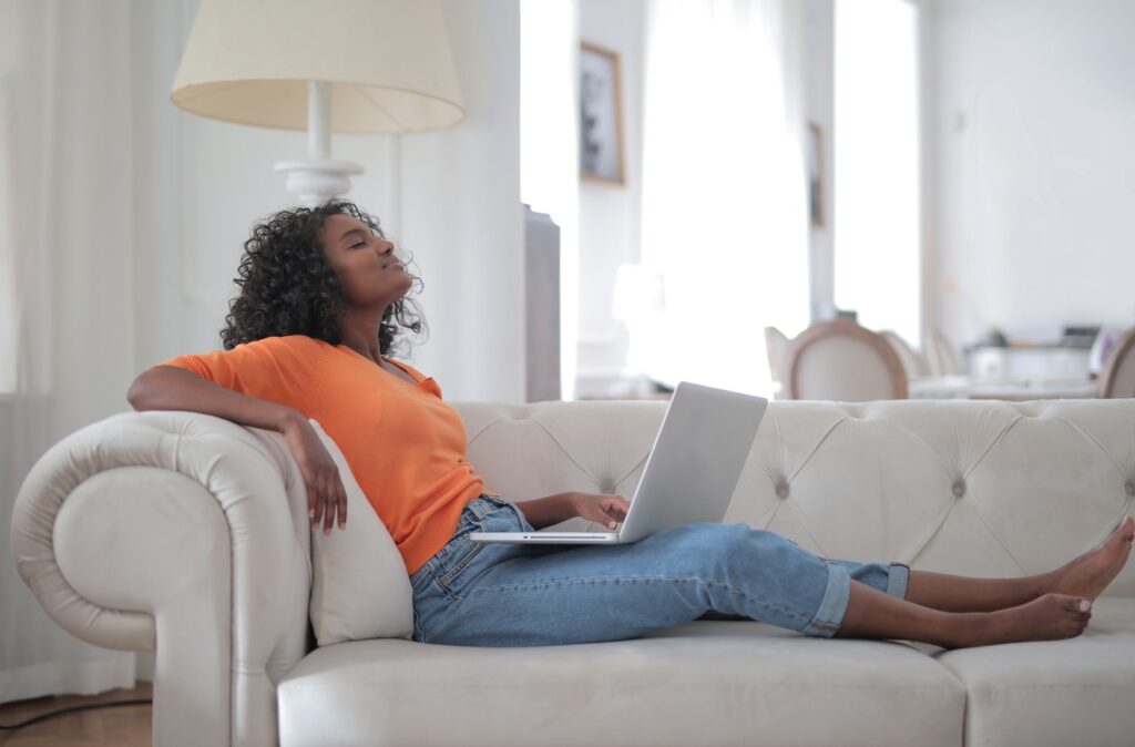 photo of a blackwoman with curly hai sitting on a white leather sofa with her head cocked back and her eyes closed. Her laptop is on her lap.