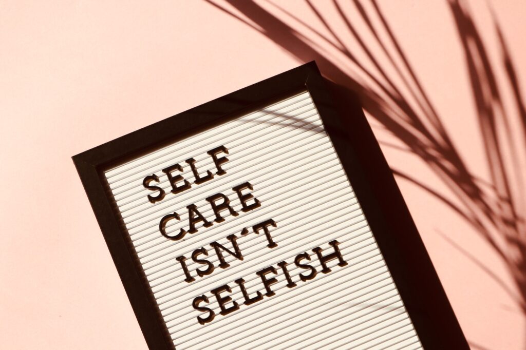 A white letterboard with the words, "self care isn't selfish" on it with a black border in the foeground to a room that has the shadow of a large fern in the back ground  in a blog about moms 