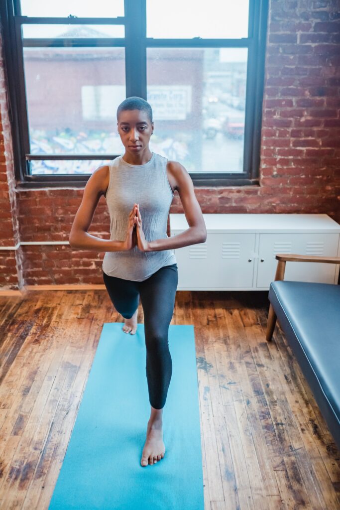 photo of a woman in a studio with recessed brick walls on a blue yoga mat doing a yoga pose. She has short cropped hair and is African American