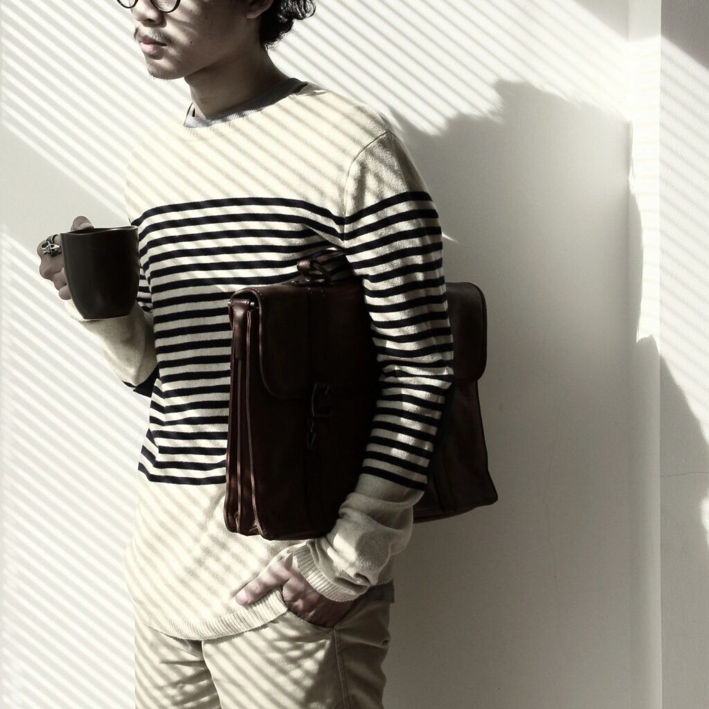 man with a brown leather briefcase under his arm and a coffee cup in another standing against wall with sunlight from blinds reflecting on his face and body.